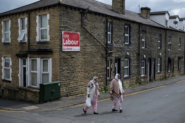 Dos mujeres cruzan una calle de la localidad de Keighley, en el noroeste de Inglaterra, cerca de un cartel que insta a votar al Partido Laborista en las elecciones, el 14 de junio de 2024 (Oli Scarff)