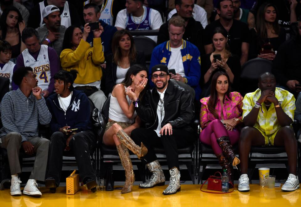 kendall jenner and bad bunny sitting courtside at a game, with kendall whispering something in his ear while laughing and covering her mouth with her hand