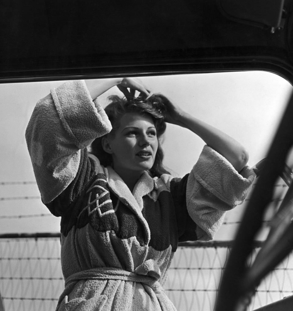 Hayworth wears a bathrobe over her swimsuit as she pins up her hair while standing next to parked car at the beach in this undated photo.