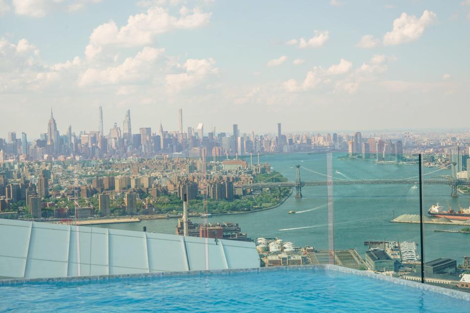 A pool in the foreground and New York City in the background with cloudy blue skies.