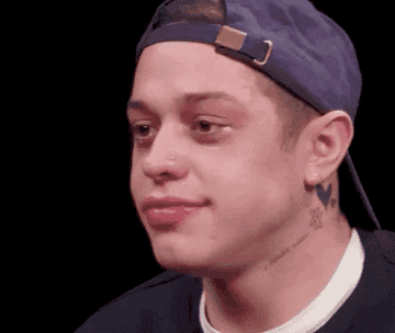 Pete Davidson wearing a blue backwards cap and a dark top, slightly smiling with a neutral expression, showing his neck tattoos