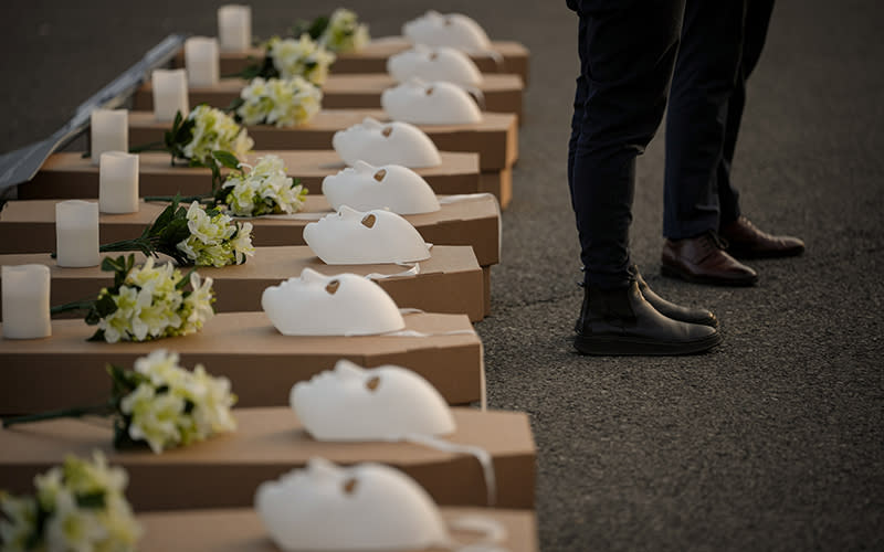 Activists stand beside a row of small mock coffins, each with a white candle, bunch of yellow flowers and a blank white mask placed on the lid.