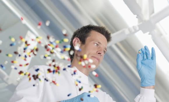 Pharmacist examining a colored tablet, with a scattering of about 50 pills on a clear glass table.