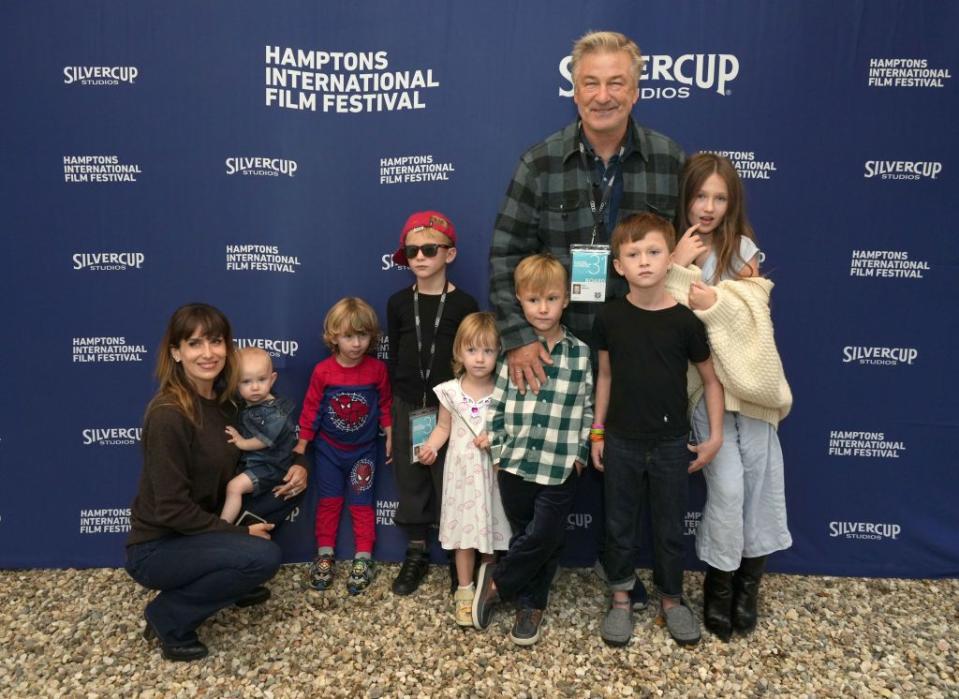 alec baldwin and his family pose for a photo in front of a blue background, his wife hilaria crouches on the left while holding their youngest child, alec smiles with standing among six other children