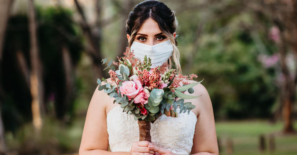A bride wearing a face mask holds a bouquet and looks at the camera