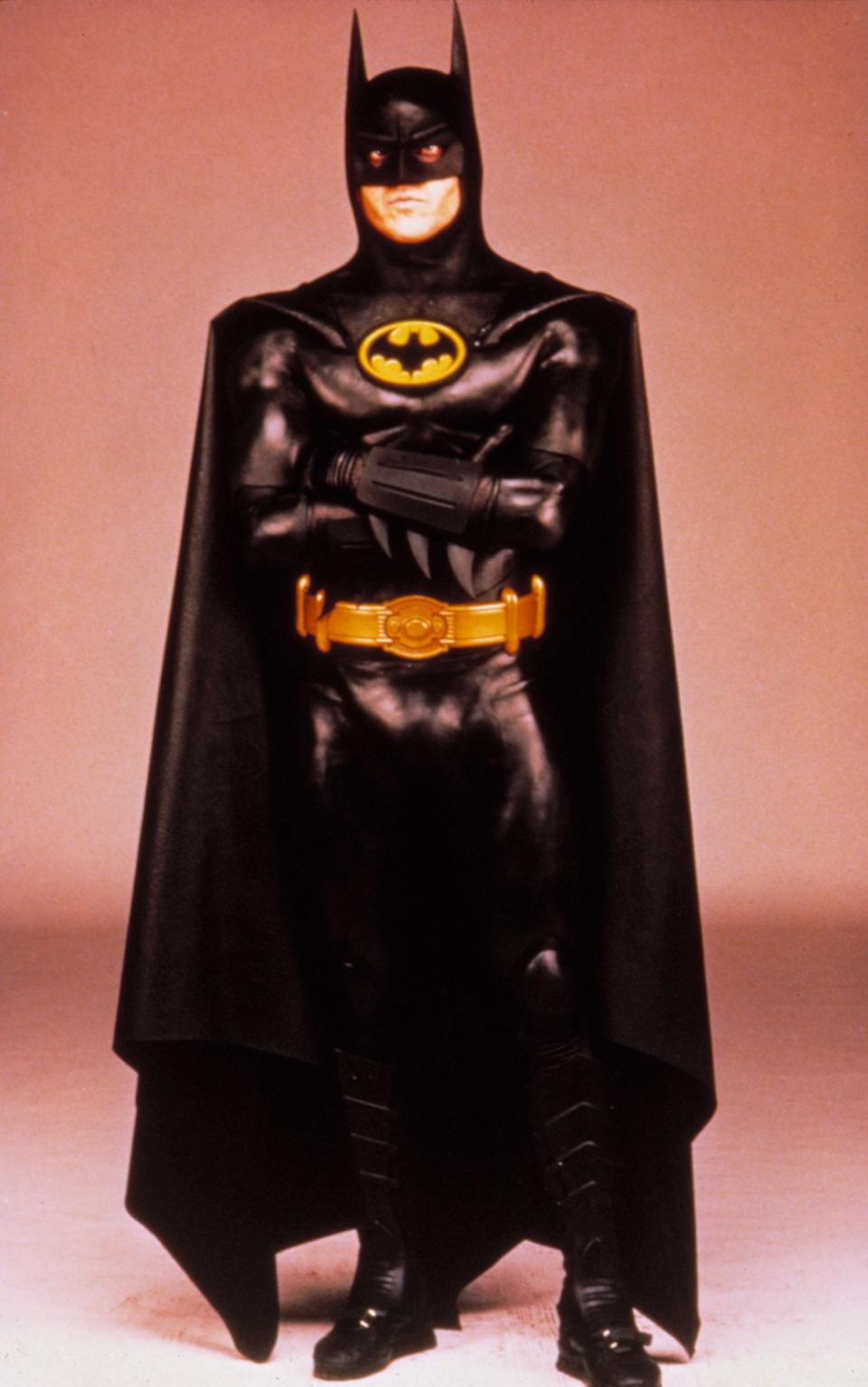 Batman stands with arms crossed in his iconic black suit, yellow utility belt, and bat symbol on his chest