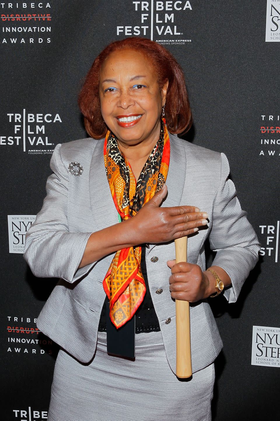 patricia bath smiles at the camera, she stands in front of a black background with white logos and wears a gray suit jacket with an orange, red, and black scarf, he holds one hand across her chest