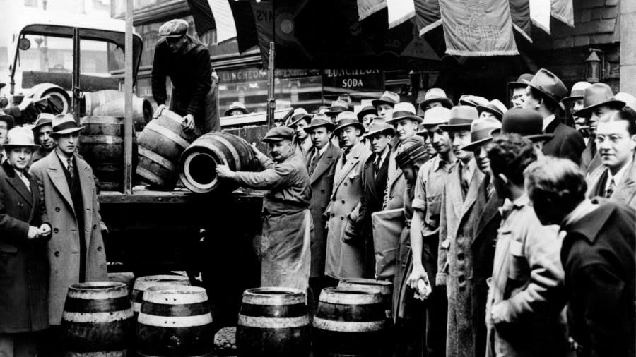 A large group of men wait outside a bar while two men work to unload barrels of beer out of a truck.