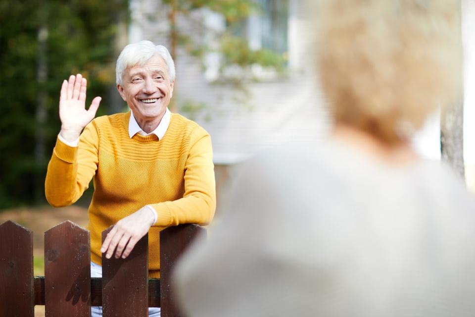 senior man in yellow sweater stands behind fence and waves at neighbor who is blurry in the foreground