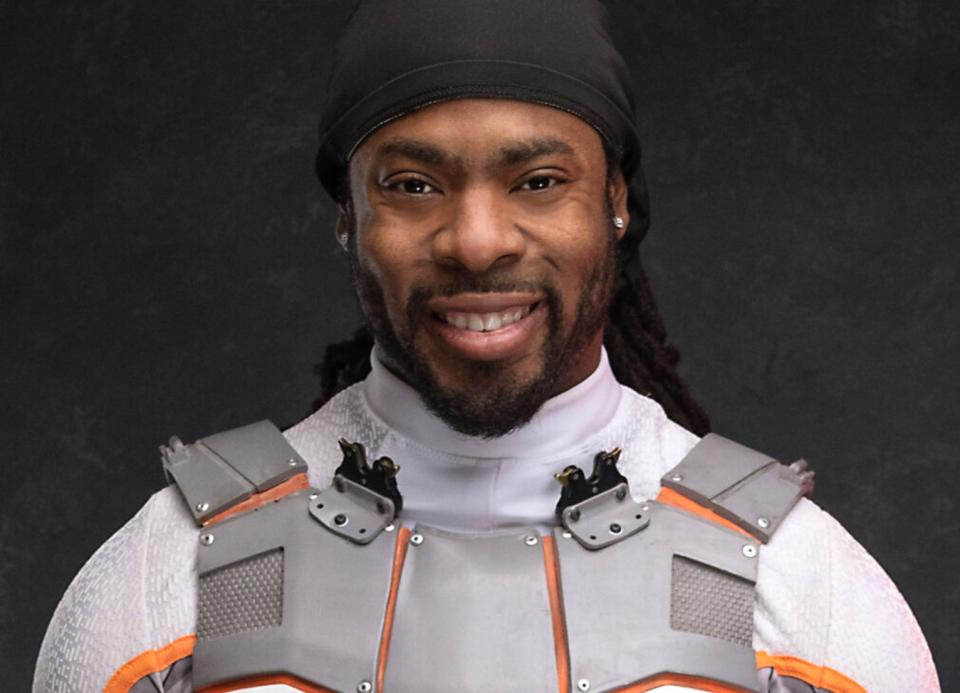 Head shot of a smiling Richard Sherman wearing a white mock turtleneck and gray plastic vest-like item.