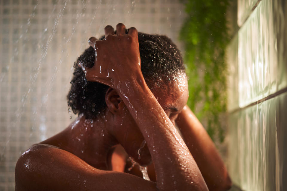 A person is taking a shower, with hands massaging wet hair.  They look relaxed and there is a green plant in the background