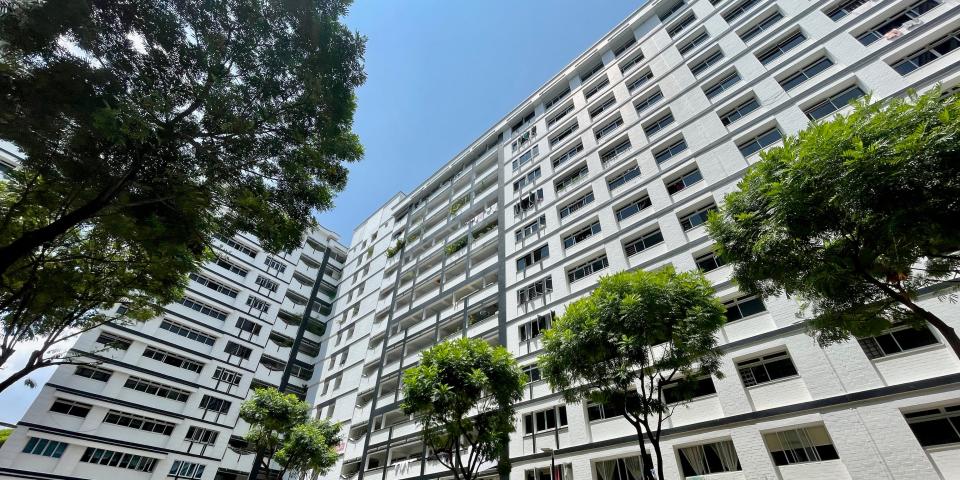 A street view of the exterior of a large apartment building with 13 floors and tall trees outside.