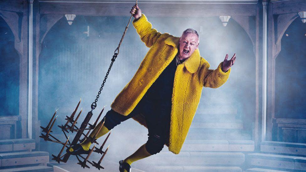 Les Dennis swinging on a chandelier, wearing a yellow furry jacket with his mouth open inside the theatre