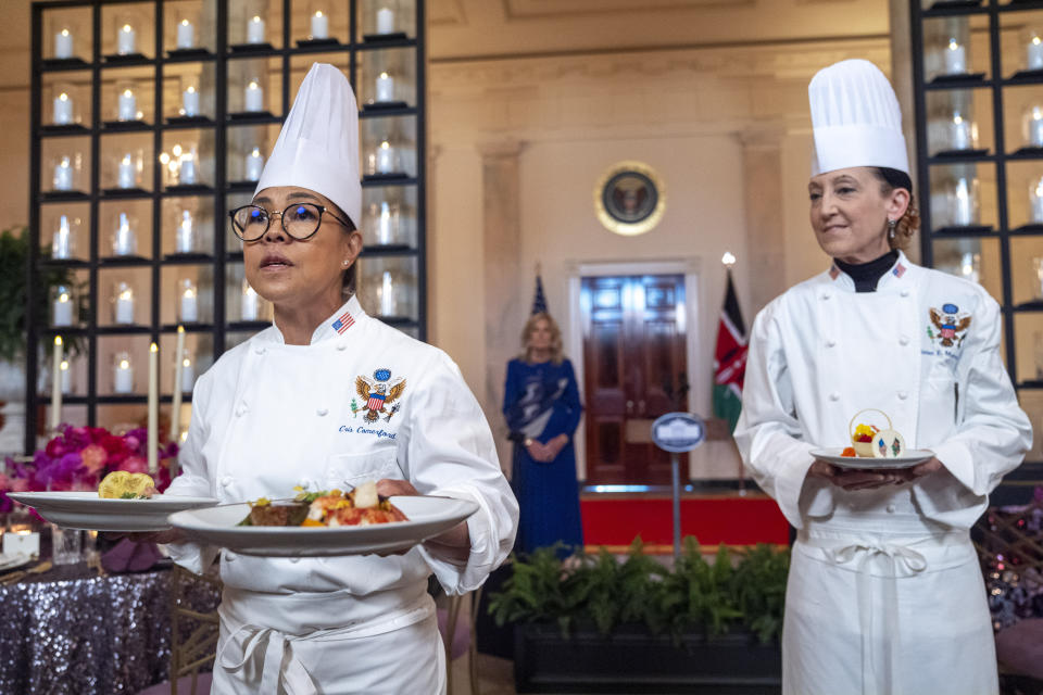 White House Executive Chef Cris Comerford, left, holds the first course, a chilled heirloom tomato soup with sourdough crisps and arbequina olive oil, and the main course of fruitwood-smoked beef short ribs, butter-poached lobster, citrus butter, baby kale, and a sweet corn purée, as White House Executive Pastry Chef Susie Morrison holds the dessert course, a white chocolate basket with banana ganache, raspberries, peaches, and candied lime zest, during a media preview, Wednesday, May 22, 2024, ahead of Thursday evening's State Dinner with Kenya's President William Ruto, at the White House in Washington. At back center is first lady Jill Biden. (AP Photo/Jacquelyn Martin)