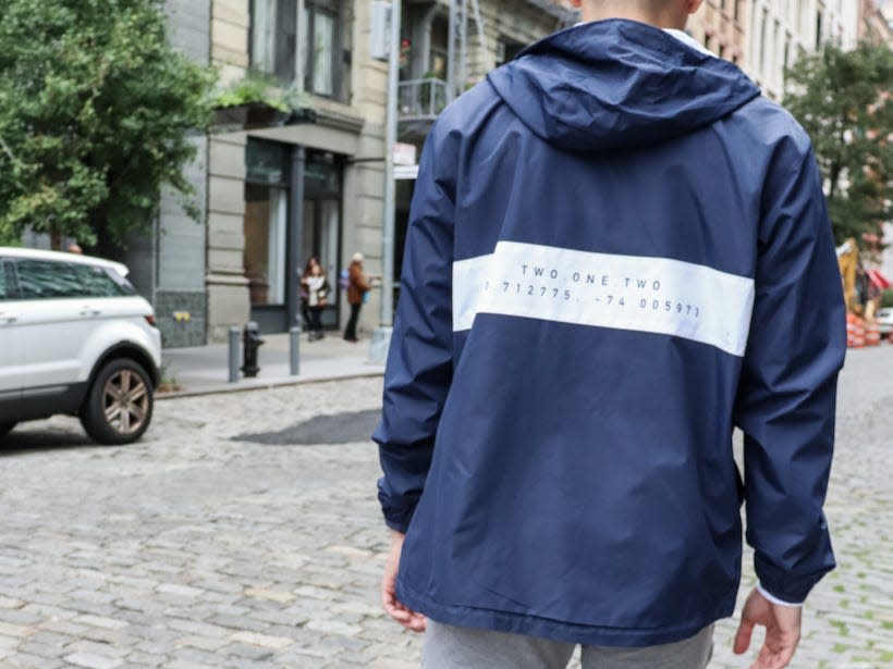 back of a man walking down a street wearing a baseball hat and a windbreaker