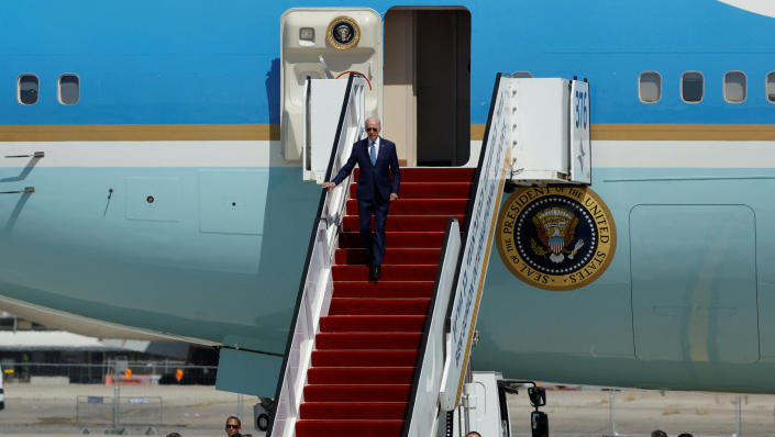 President Biden, in sunglasses, descends red-carpeted stairs, with the presidential seal visible on the plane.