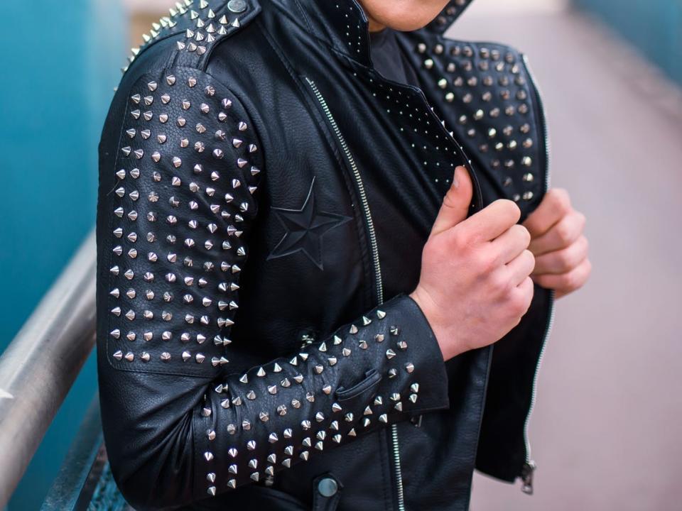 A young man wears a punk-style studded black leather jacket while posing on a ramp in front of a teal building exterior.