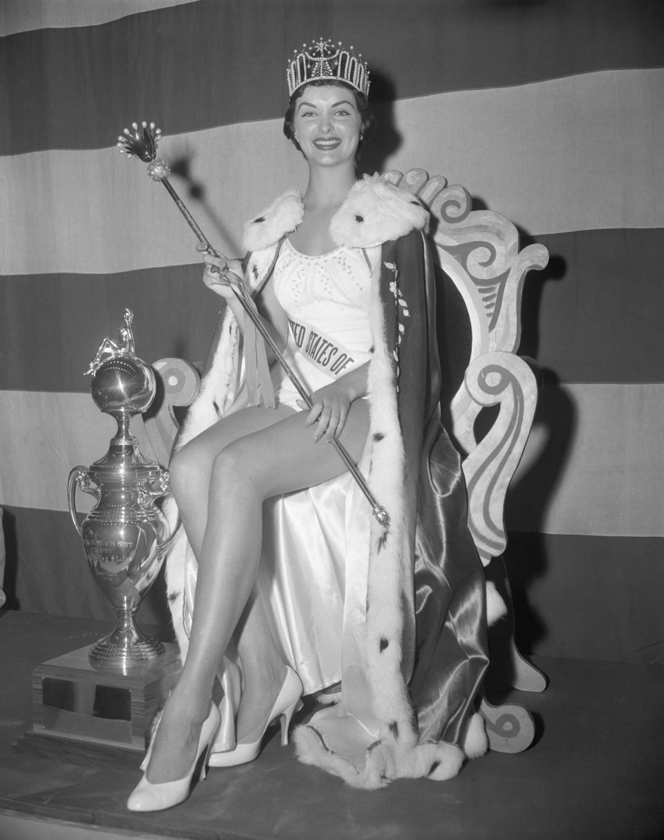 Miss USA 1957 Leona Gage sits on a throne with a tiara, scepter, and robe.