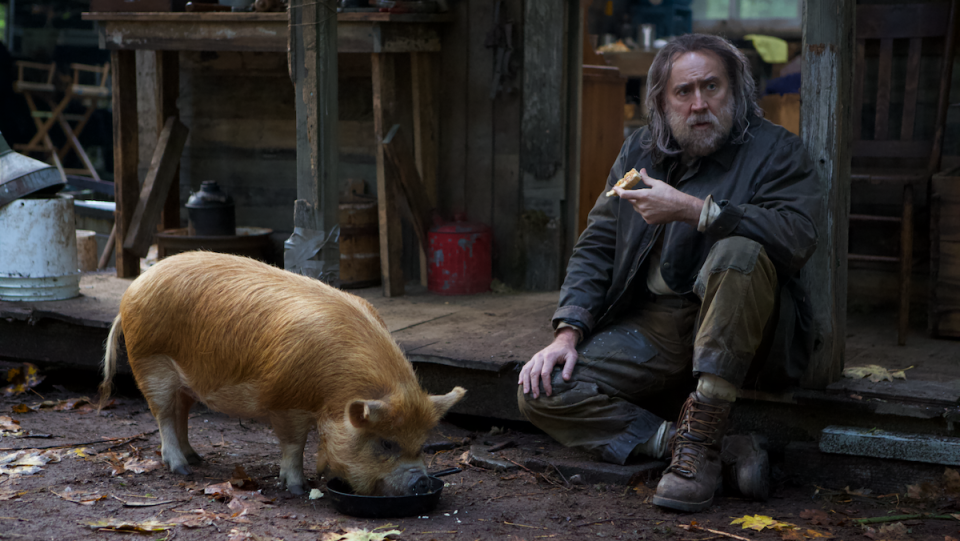 A long-haired Nicolas Cage sits on the ground with a pig.