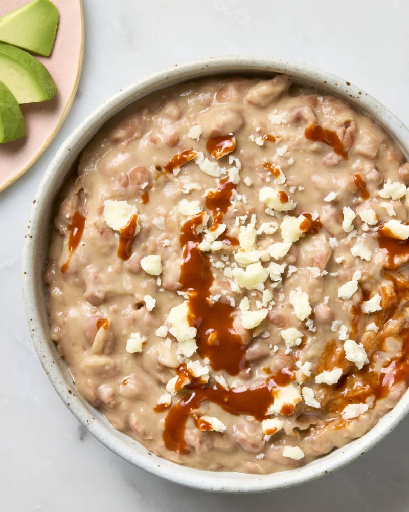 An overhead view of a bowl of refried beans with cotija cheese and hot sauce, with sliced of avocado on the side.