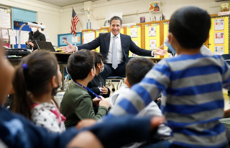 A group of kids sits on the floor in front of a man in a suit