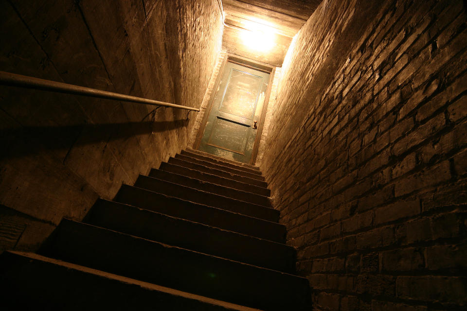 A dimly lit, narrow staircase made of stone leads up to a closed door, creating a mysterious and slightly eerie atmosphere