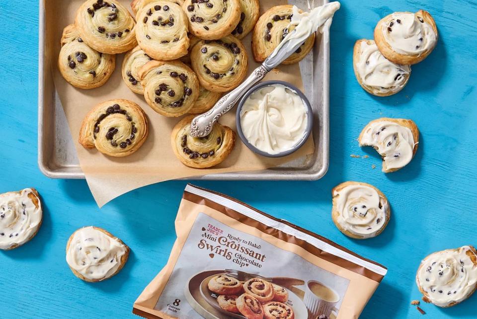 trader joe's ready to bake mini croissant swirls stacked in baking pan next to crock of cream cheese frosting some swirls frosted, one has a bite