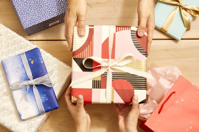 Overhead shot of someone handing over a gift wrapped in red, pink and gold abstract paper, to someone grabbing it with two hands.  On the light wood surface theres an assortment of other wrapped gifts and gift bags.