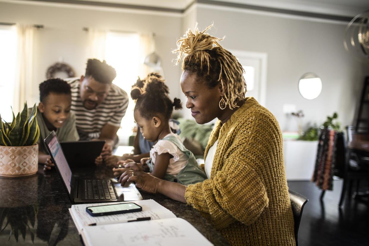 Mother working from home while holding toddler, family in background