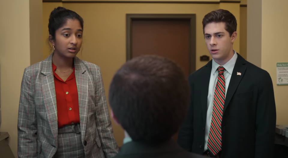 Maitreyi Ramakrishnan stands next to Darren Barnet in an office hallway, looking at a person with their back to the camera