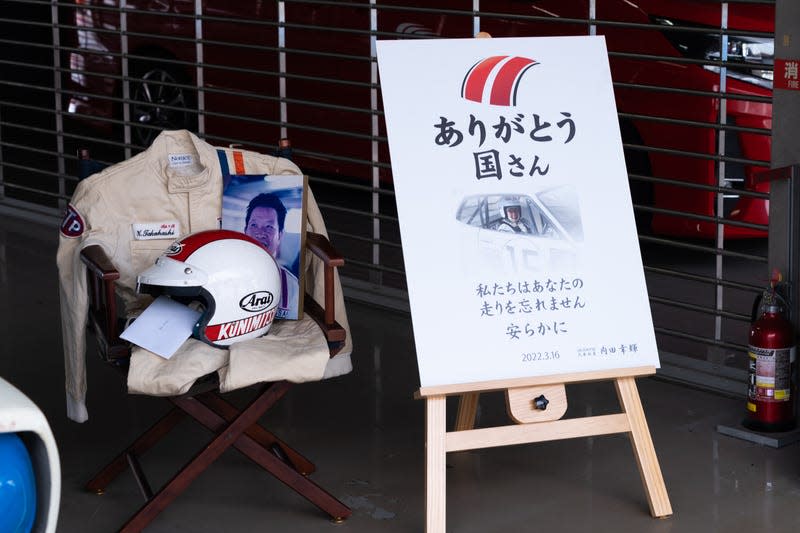 a helmet and racing jacket sit on a chair next to a poster honoring the recently-deceased japanese racing driver Kunimitsu Takahashi