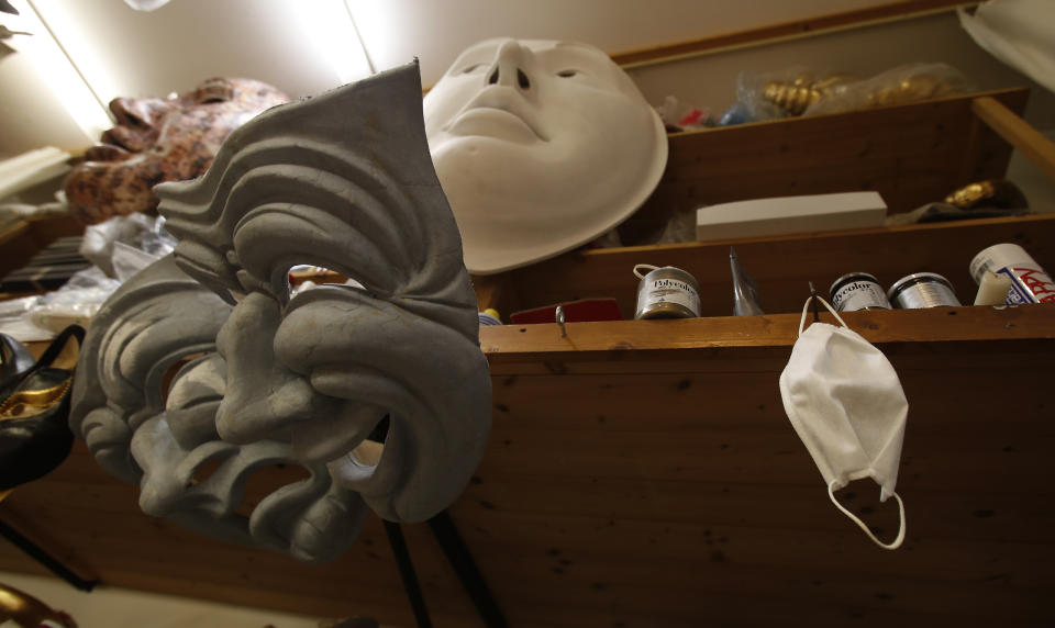 A sanitary face mask hangs next to carnival mask in an artisan workshop in Venice, Italy, Saturday, Jan. 30, 2021. In another year, masks would be an accepted sign of gaiety in Venice, an accessory worn for games, parties and crowds. Since the onset of the COVID-19 pandemic face masks are worn now to protect, not amuse. (AP Photo/Antonio Calanni)