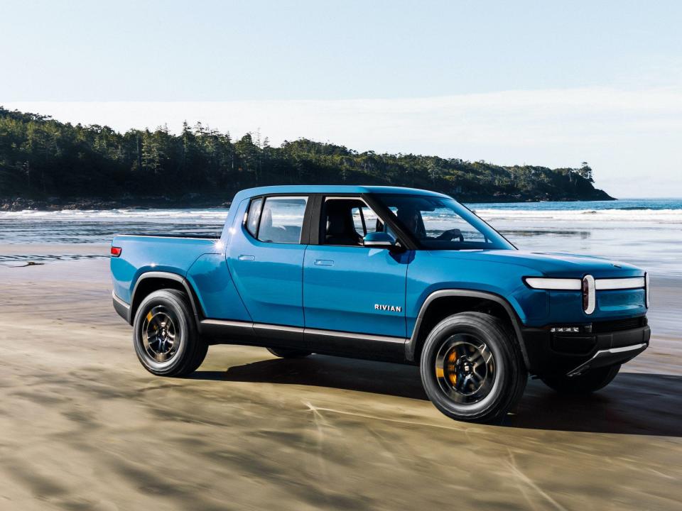 A bright blue pickup truck parked on a beach in front of a body of water with some distant greenery in the background.