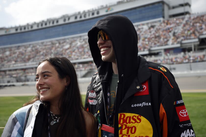 Pete Davidson and Chase Sui smile, facing to the left, waiting at Daytona International Speedway in Daytona Beach, Florida
