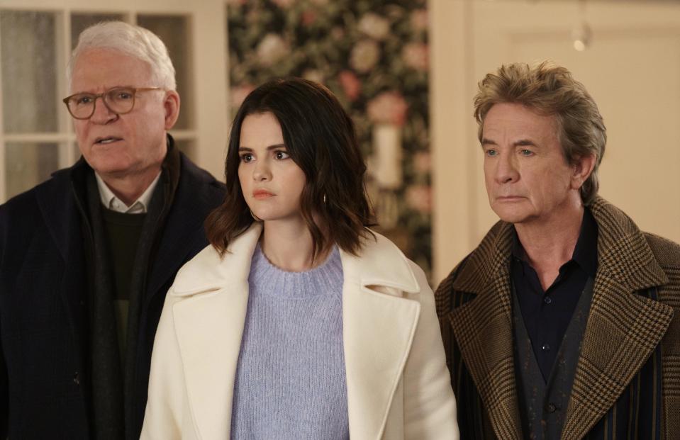 Steve Martin, Selena Gomez, and Martin Short stand together in a room, looking serious. Selena is wearing a white coat over a lavender top