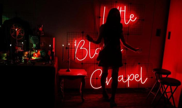 An attendee poses for a photograph inside the Little Black Chapel at The Satanic Temple&#39;s Satancon 2023. The figure is silhouetted against a wall lit by a red neon sign reading &#39;Little Black Chapel&#39;. A table with candles on it stands to the left.