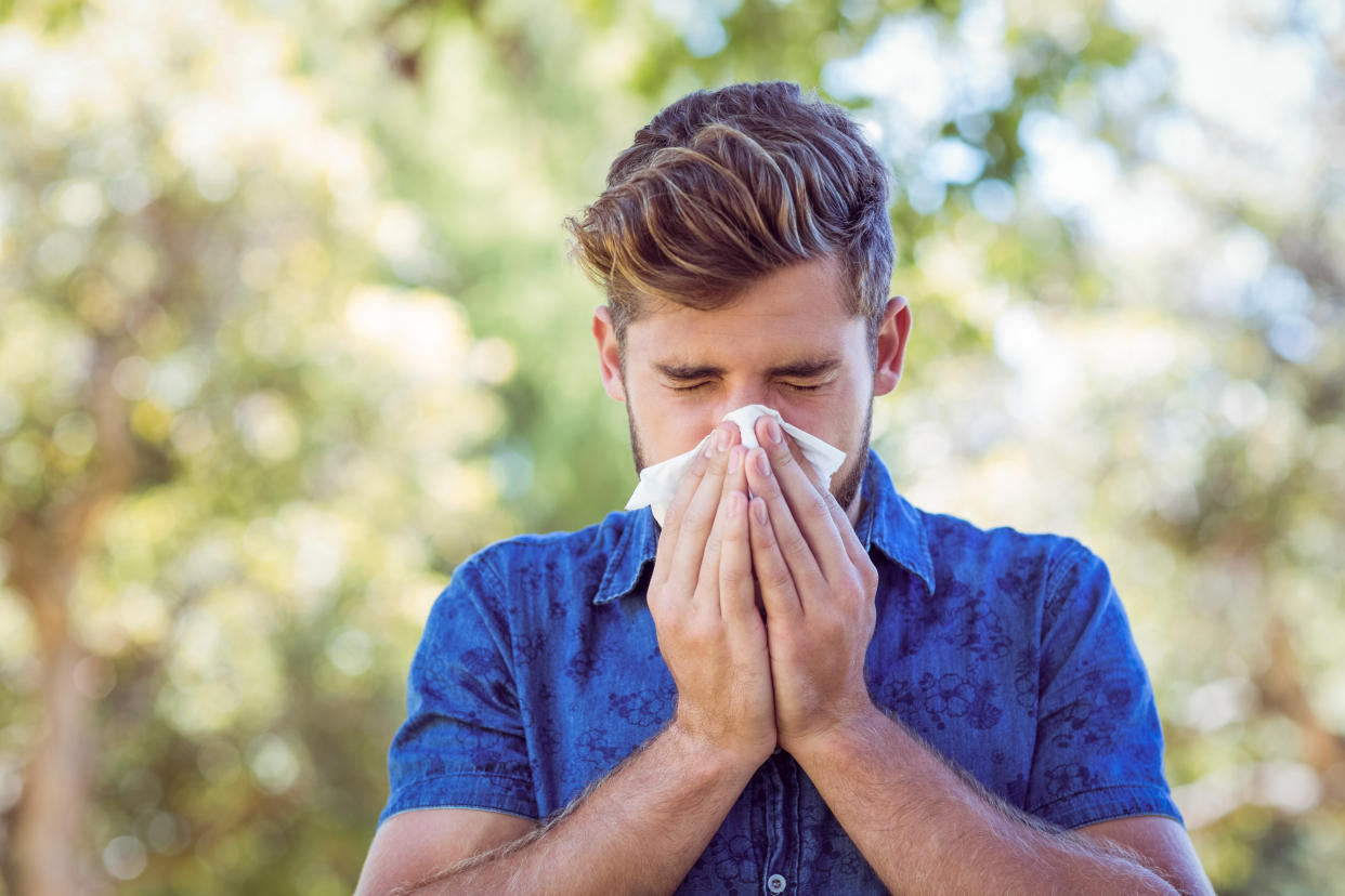 A man wearing a blue, collared short-sleeve shirt and sporting a pompadour blows his nose into a tissue using two hands.