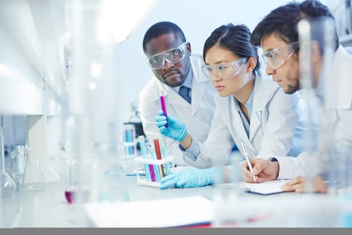 Three scientists in a lab, with one holding a test tube and another writing on a notepad