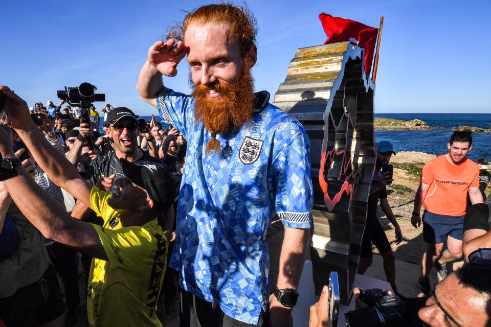 TOPSHOT - British runner Russ Cook gestures as he poses for a picture with the memorial sign marking the northern-most point of Africa upon arrival at Cape Angela, northeast of Tunis, on April 7, 2024 while surrounded by supporters who joined him for the final leg of the 16,000 kilometre challenge to run across the African continent from South Africa's Cape Agulhas to Tunisia's Cape Angela to raise money for charity. (Photo by FETHI BELAID / AFP) (Photo by FETHI BELAID/AFP via Getty Images)