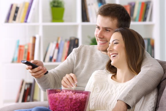 A smiling young couple snuggles up on the couch, TV remote in hand.