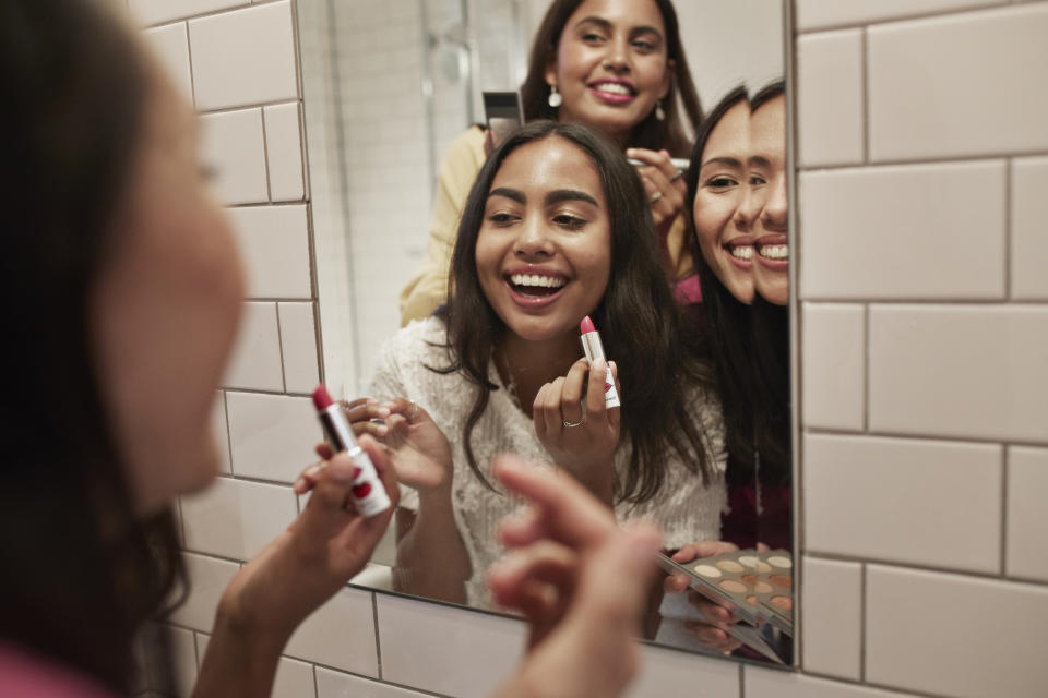 Three friends are smiling and applying makeup in front of a mirror