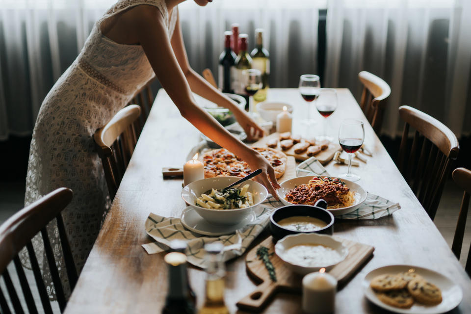 A person in semi-formal attire arranges food on a dining table set with various dishes, candles, and wine glasses