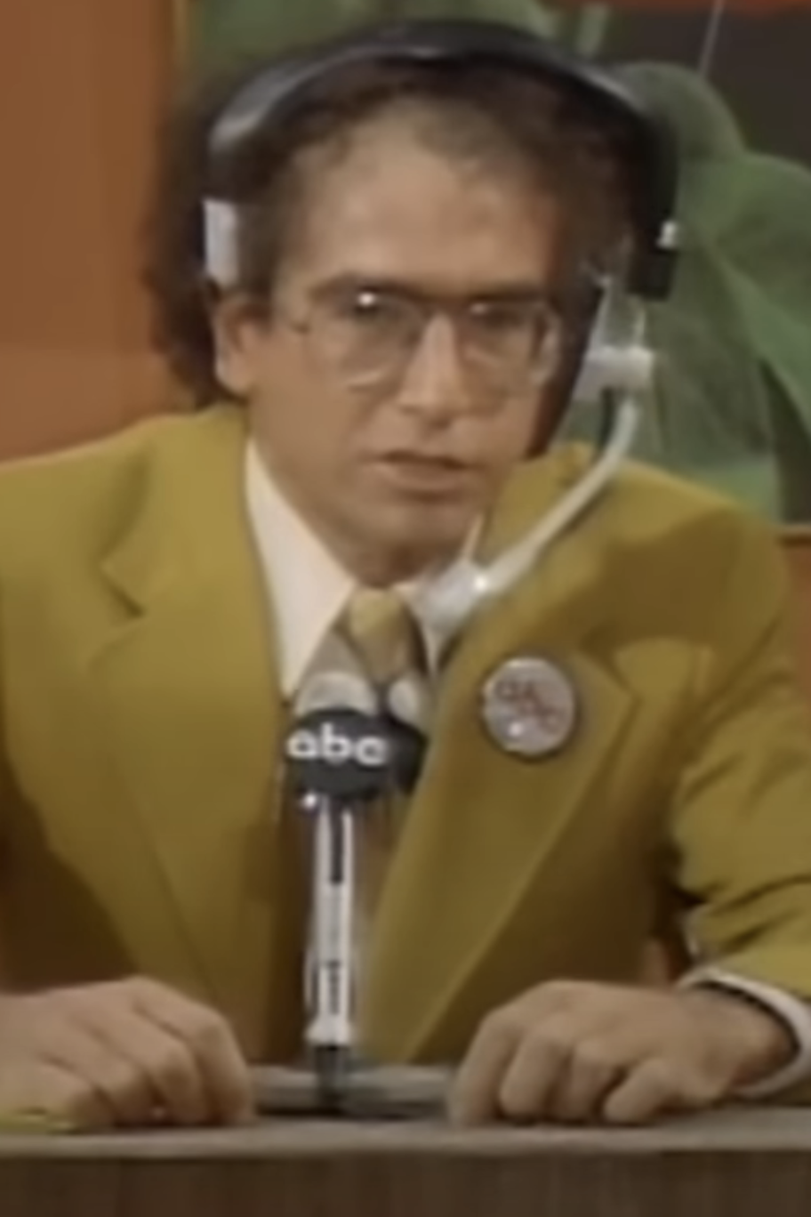 Larry David wearing a yellow suit, glasses, and a headset, seated at a desk with an ABC microphone, appears to be engaged in a serious discussion