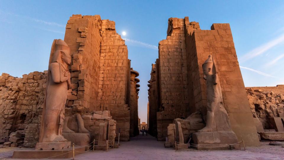 tall egyptian ruins with tall figures and walls with a blue sky and sun in the background.