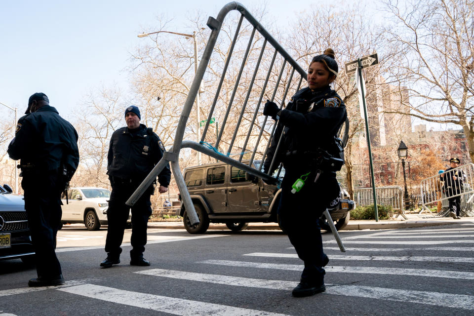 A female police officer lifts a section of barricade across a crosswalk, with a park in the background.