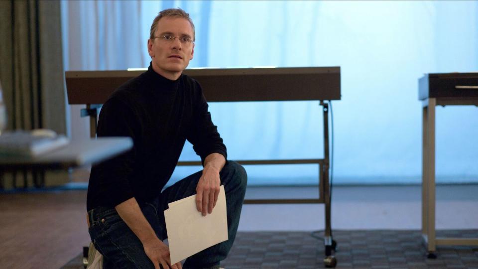 michael fassbender dressed as steve jobs crouches on a rug while holding a piece of paper in his hand and looking toward the camera, he has on a black turtleneck and black pants with white sneakers and wire rimmed glasses