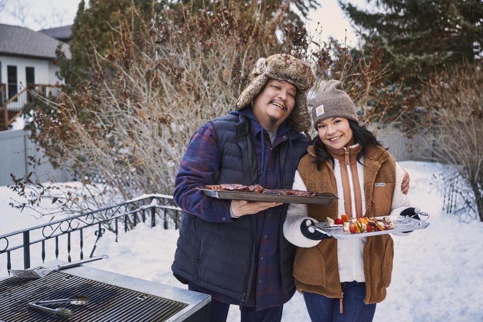 A couple grilling outside during winter hold up their BBQ while outfitted in winter vests, fleece sweaters and winter hats.