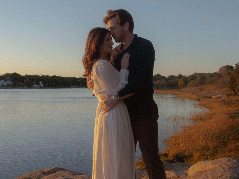 Anna, wearing a flowy white dress, embraces Trevor on the shoreline in New England.
