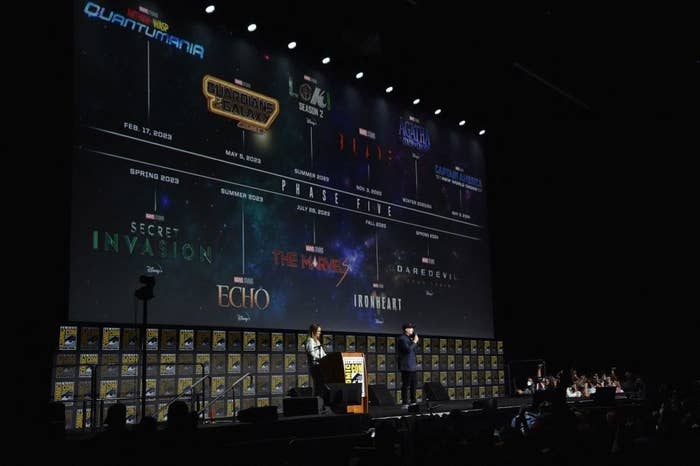 Two people on stage at Comic-Con with the timeline of Marvel's Phase Five on a giant screen behind them