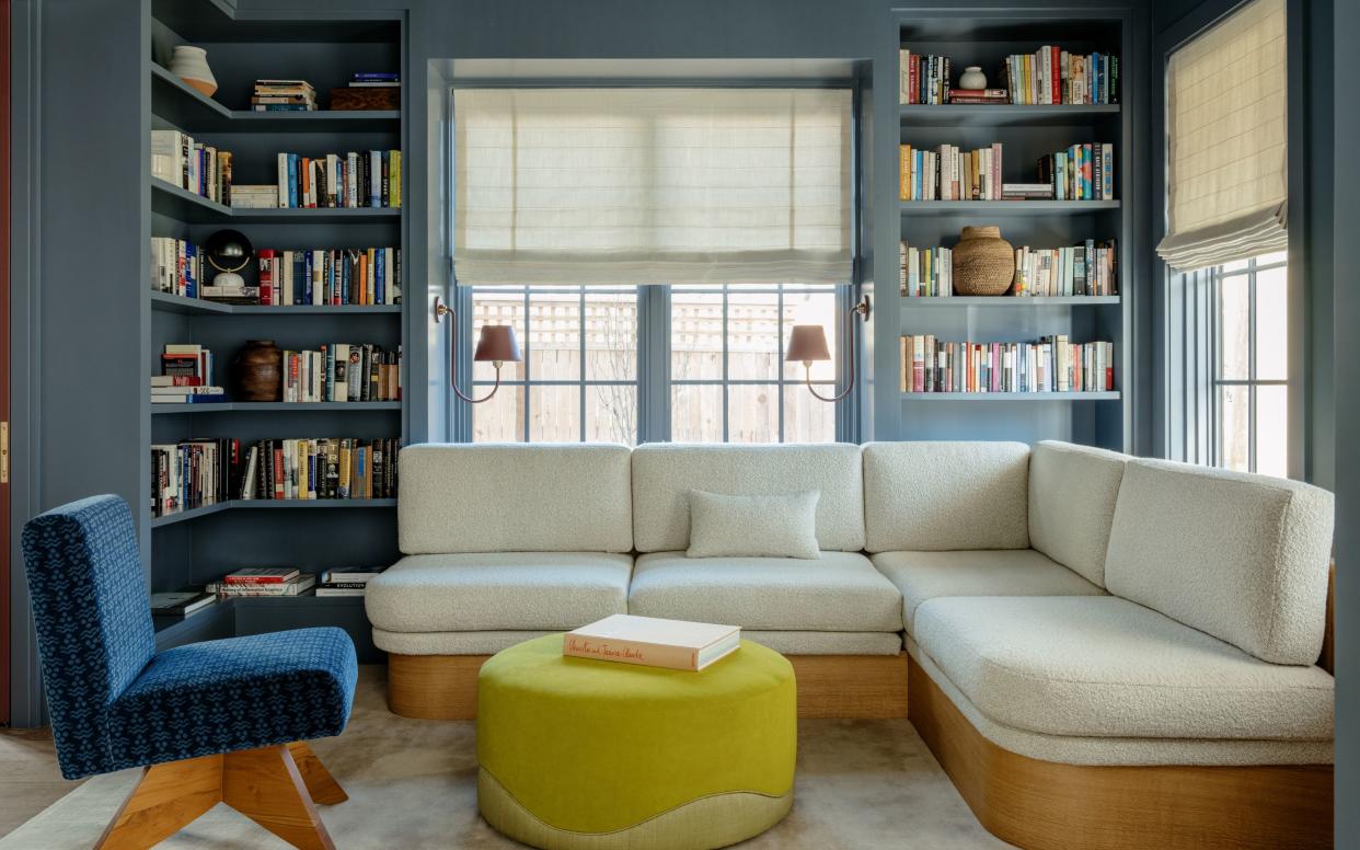  Home library snug with blue walls and shelvesm light colored sofa, blue accent chair, and yellow pouffe. 